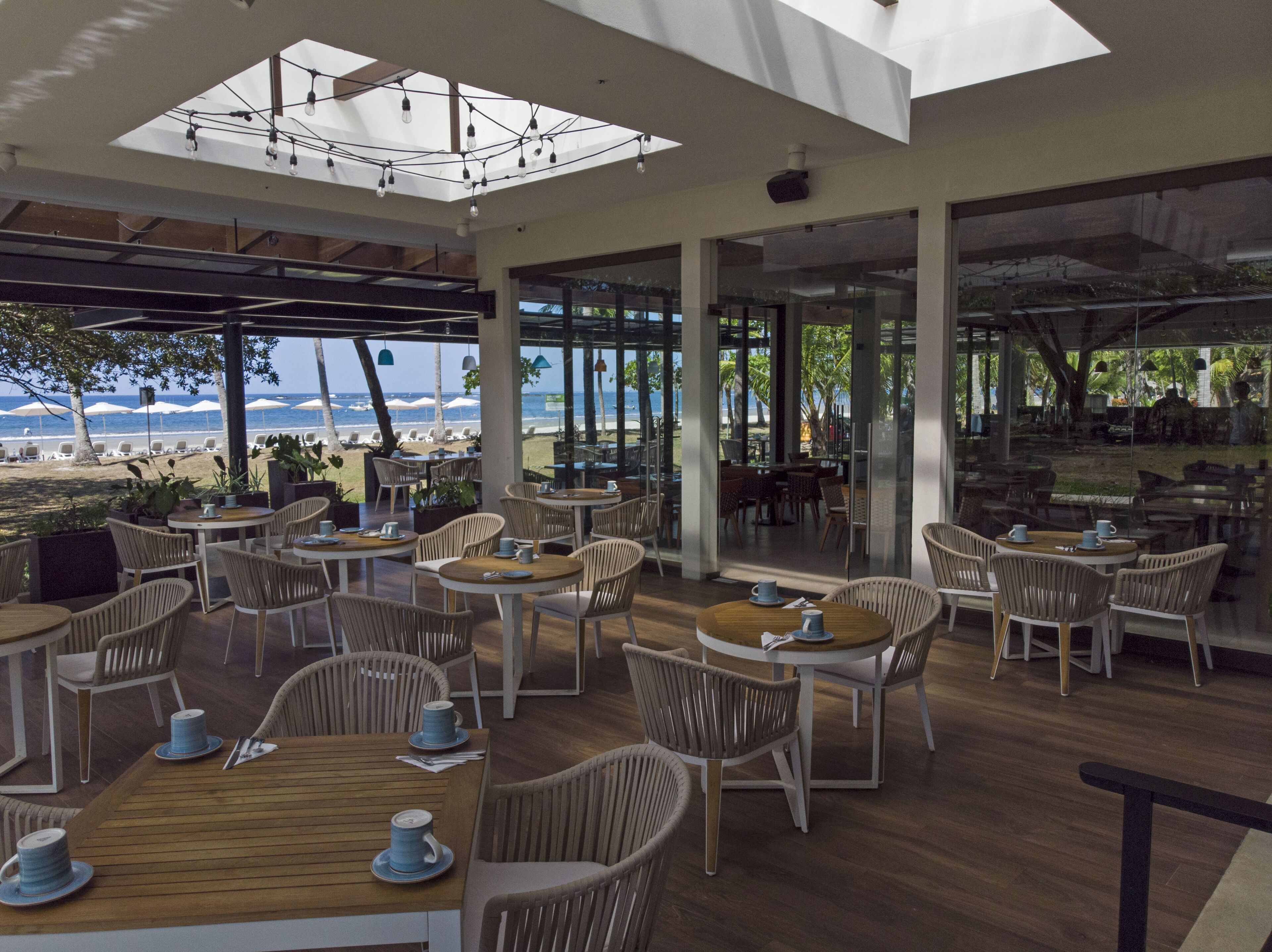 A spacious outdoor dining area with multiple tables and chairs, large windows offering views of the beach and ocean, and a modern architectural design with a skylight.