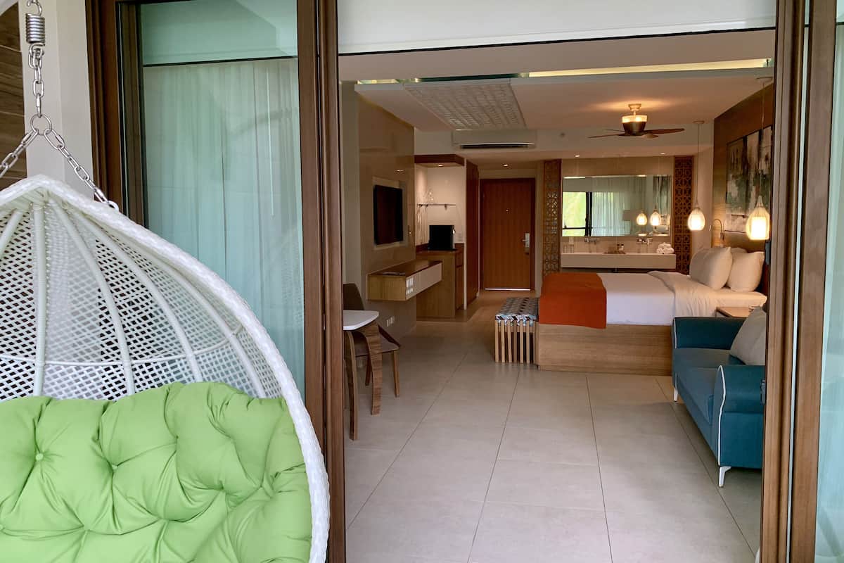 A hotel room with a green cushioned chair, a blue sofa, a small table, a desk with a computer, and a bathroom with a mirror and sink.