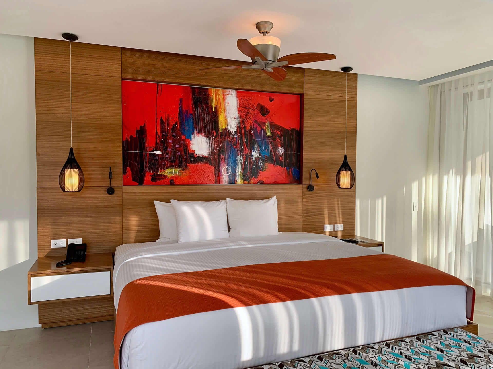 Una habitación de hotel con una cama grande, un ventilador de techo, un cuadro con un paisaje urbano y dos lámparas de noche.