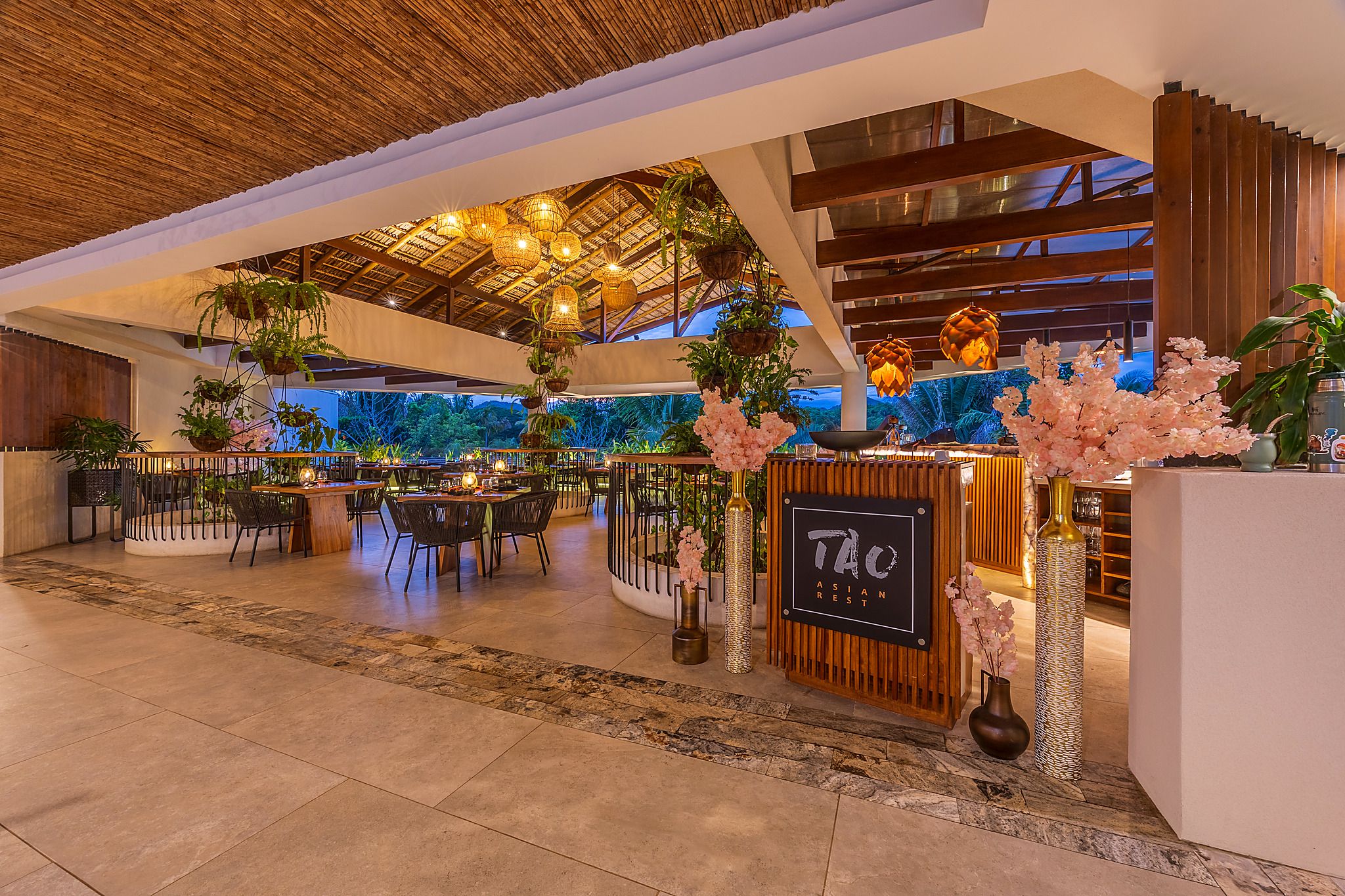 A spacious indoor area with multiple dining tables and chairs, a reception desk, and hanging plants.