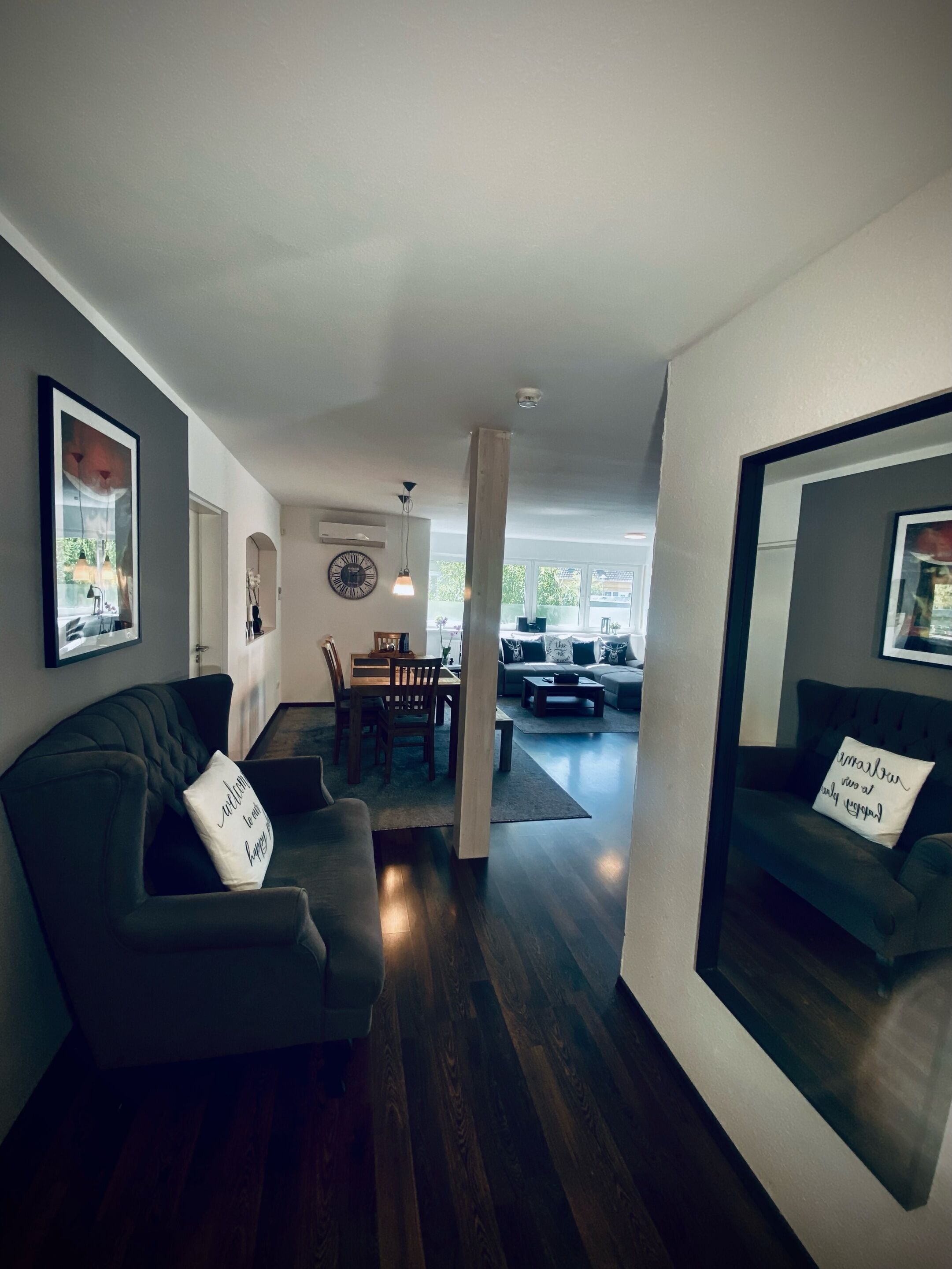 A modern living room with a dark grey sofa, a dining area with a wooden table and chairs, and a large window providing natural light.
