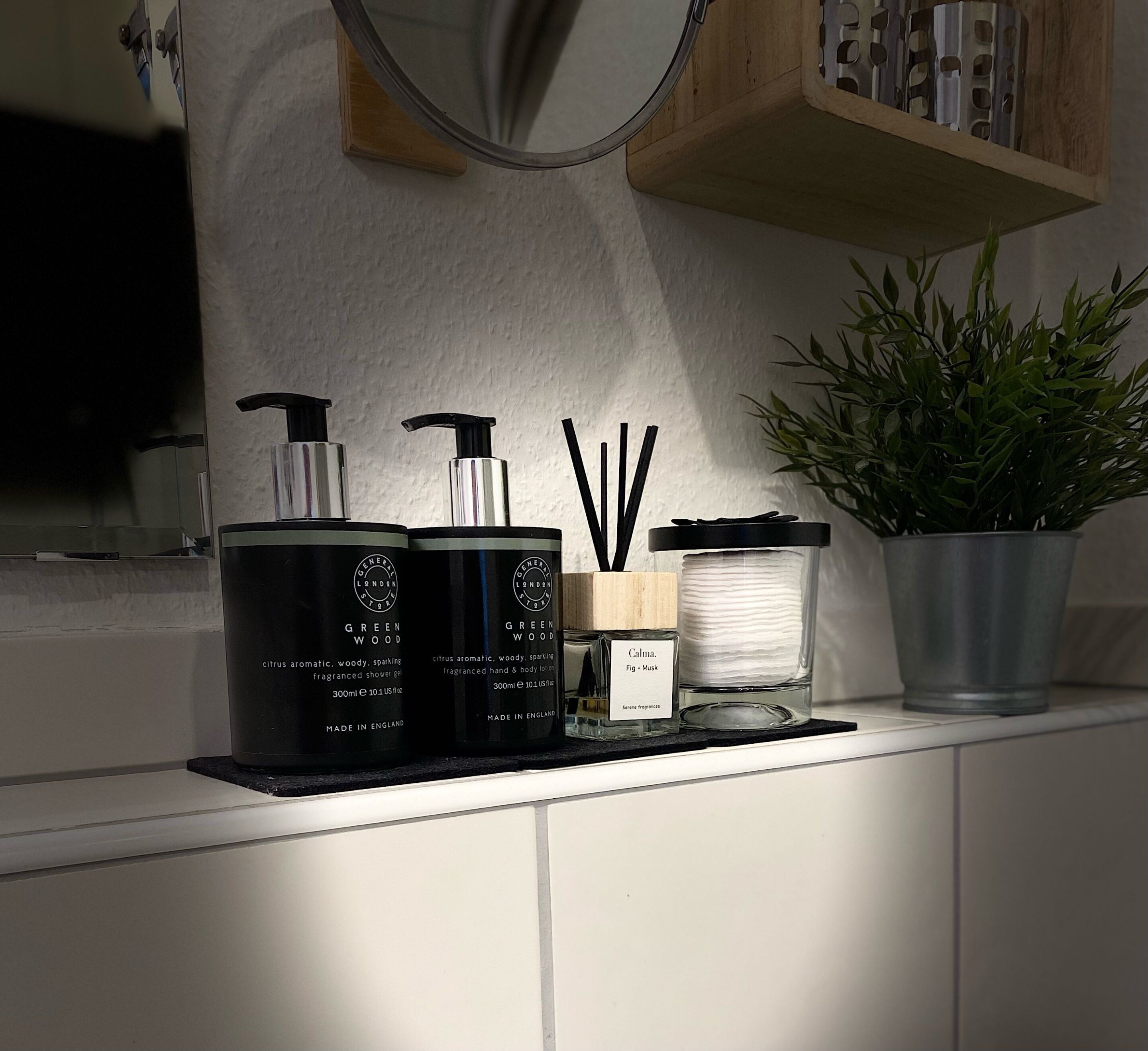 A bathroom shelf with toiletries including a green wood scented shower gel, a hand and body lotion, and a small potted plant.