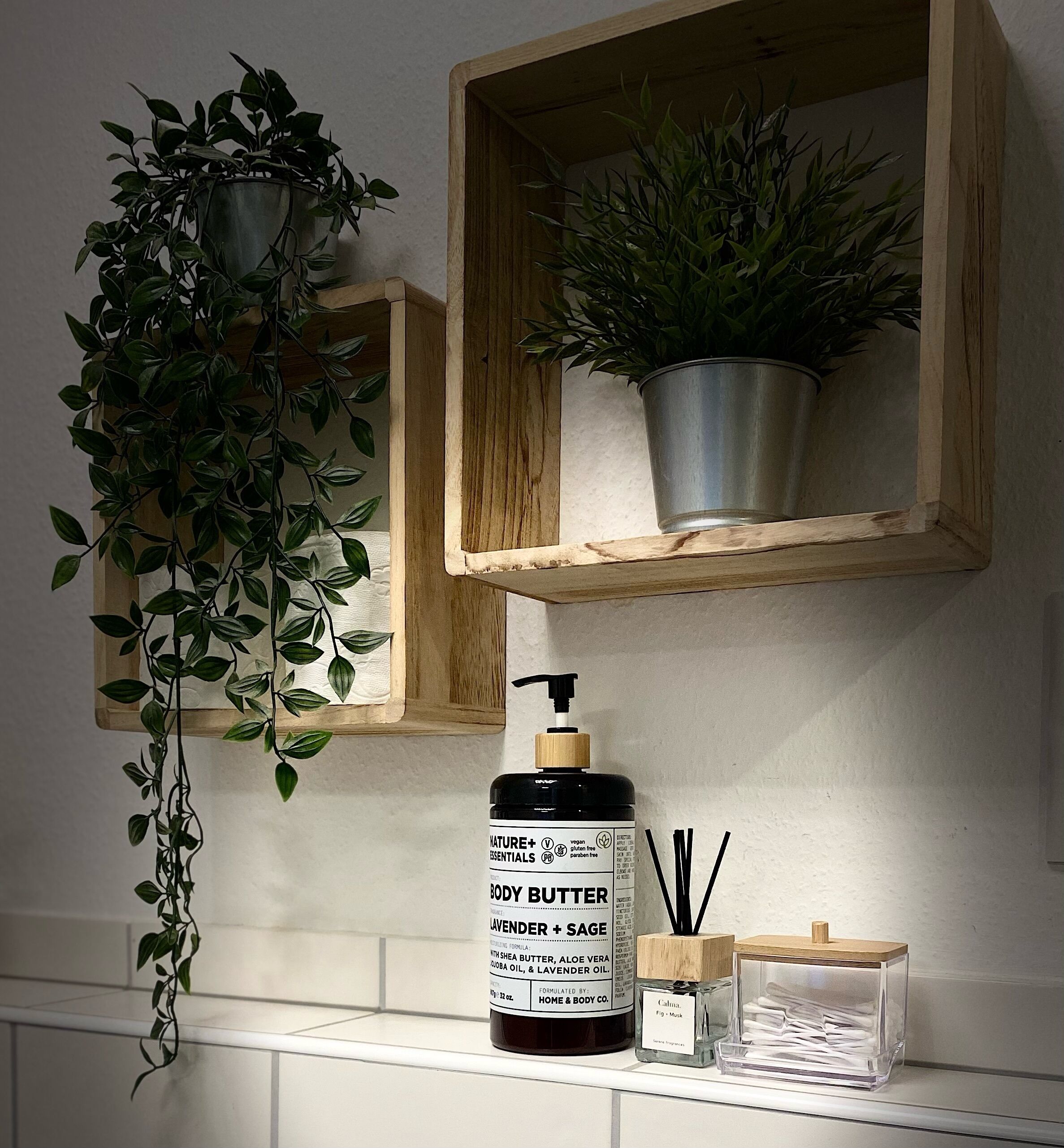 Ein Badezimmerschrank mit einer Bodybutter-Flasche, einem kleinen Behälter für Reed-Diffuser und zwei Blumentöpfen.