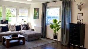 A living room with a sofa, coffee table, and a plant. There is a window with curtains and a wall-mounted deer head decoration.
