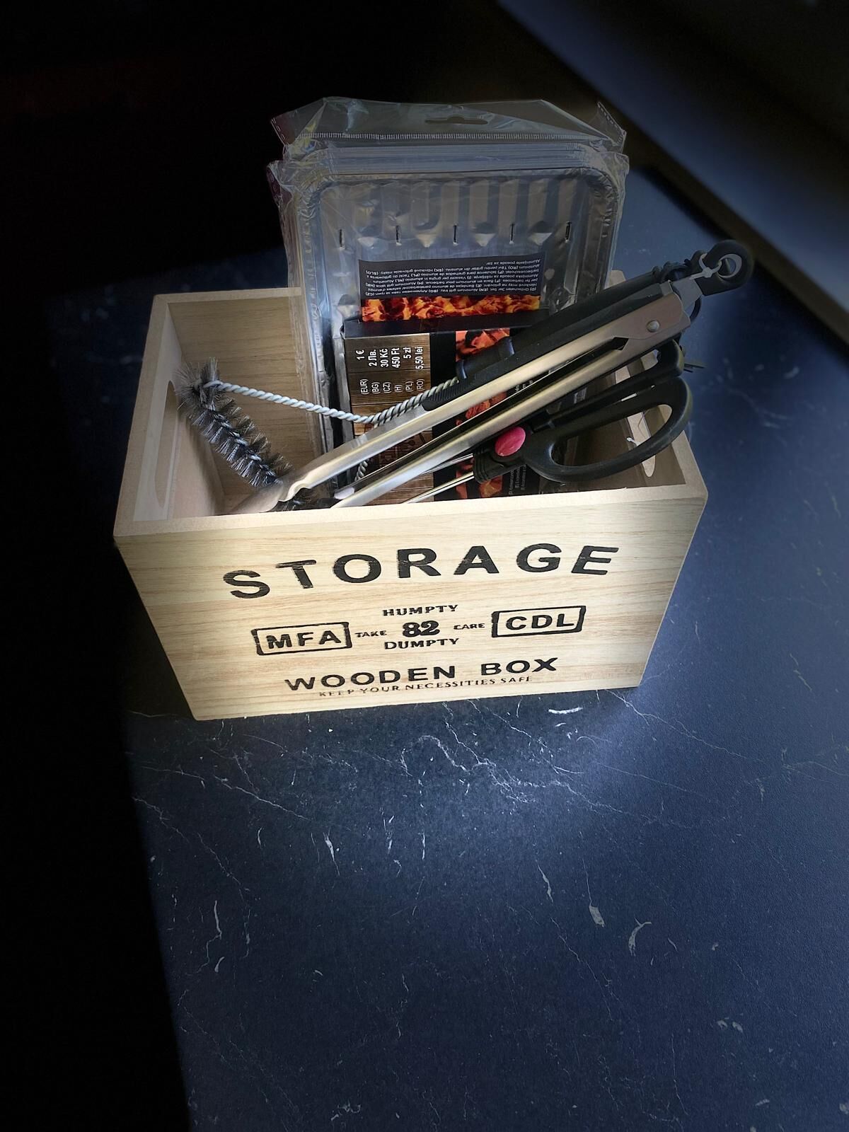 Una caja de madera con la etiqueta 'STORAGE' y compartimentos para artículos 'HUMPTY' y 'CDL', que contiene diversas herramientas y un paquete.