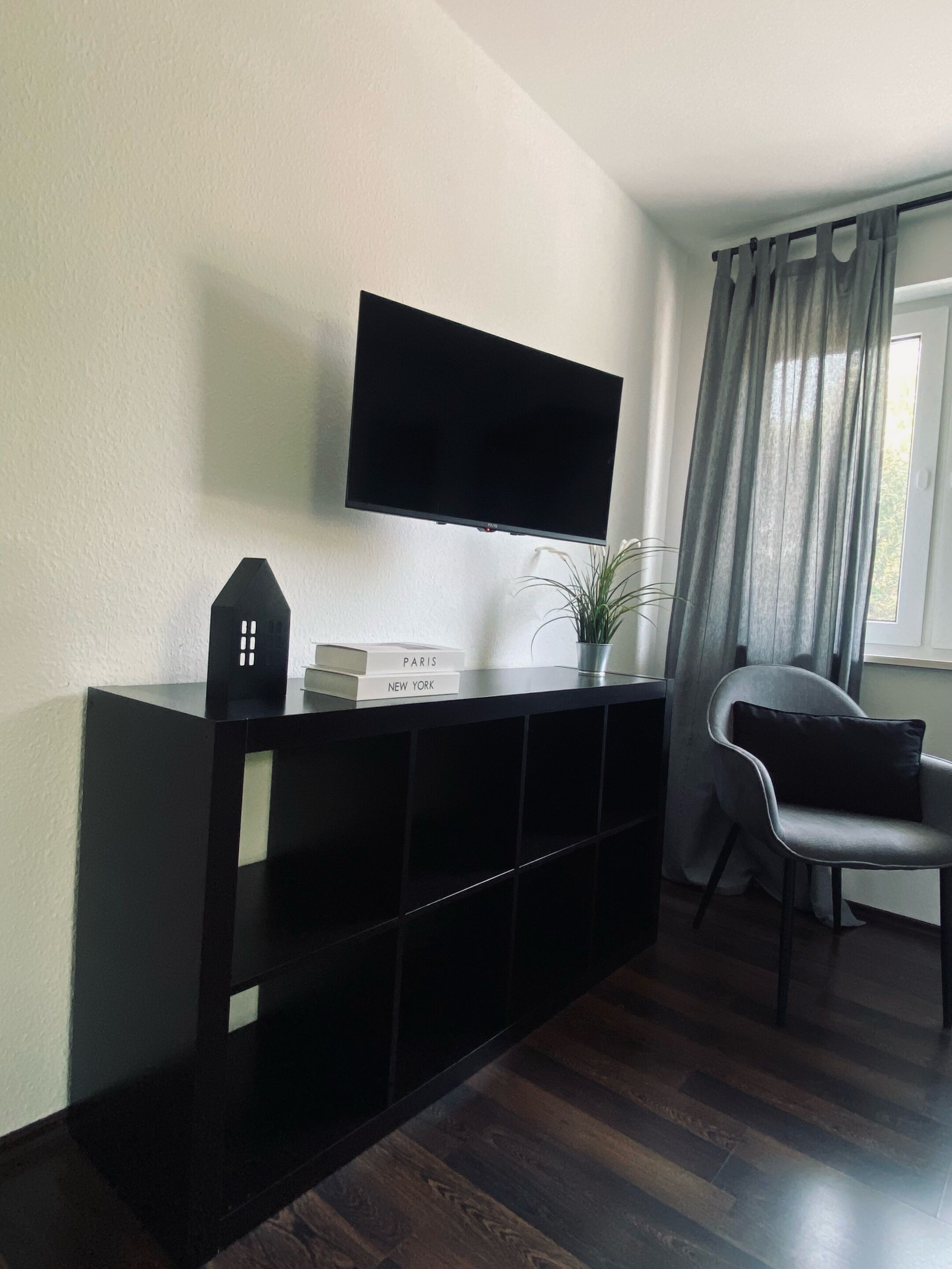 A modern living room with a flat-screen TV mounted on the wall, a black entertainment unit, a grey chair, and a window with curtains.