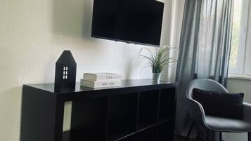 A modern living room with a flat-screen TV mounted on the wall, a black entertainment unit, a grey chair, and a window with curtains.