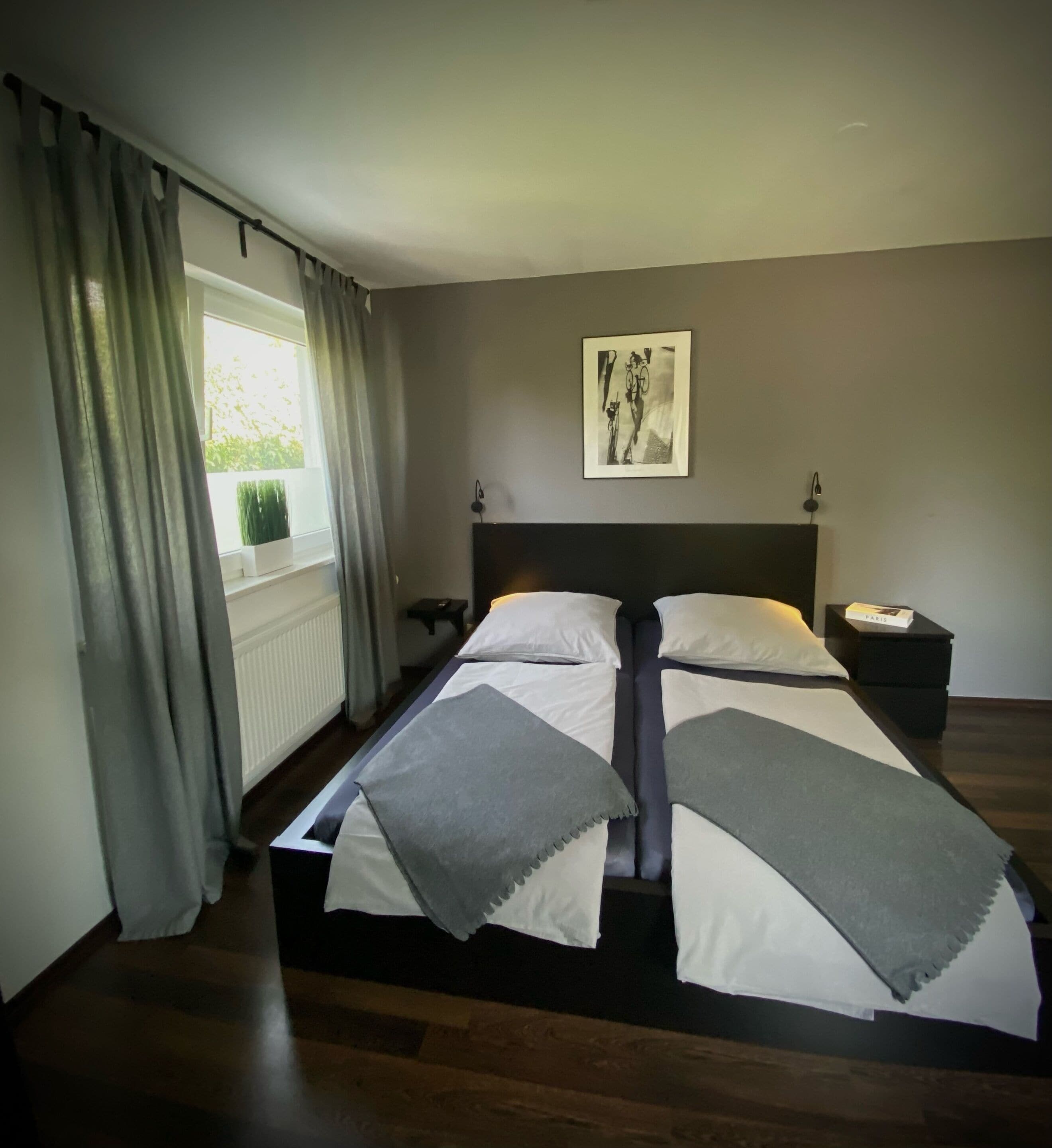 Ein Schlafzimmer mit Bett, einem Fenster mit Vorhängen, einem Wandbild und einem Nachttisch.