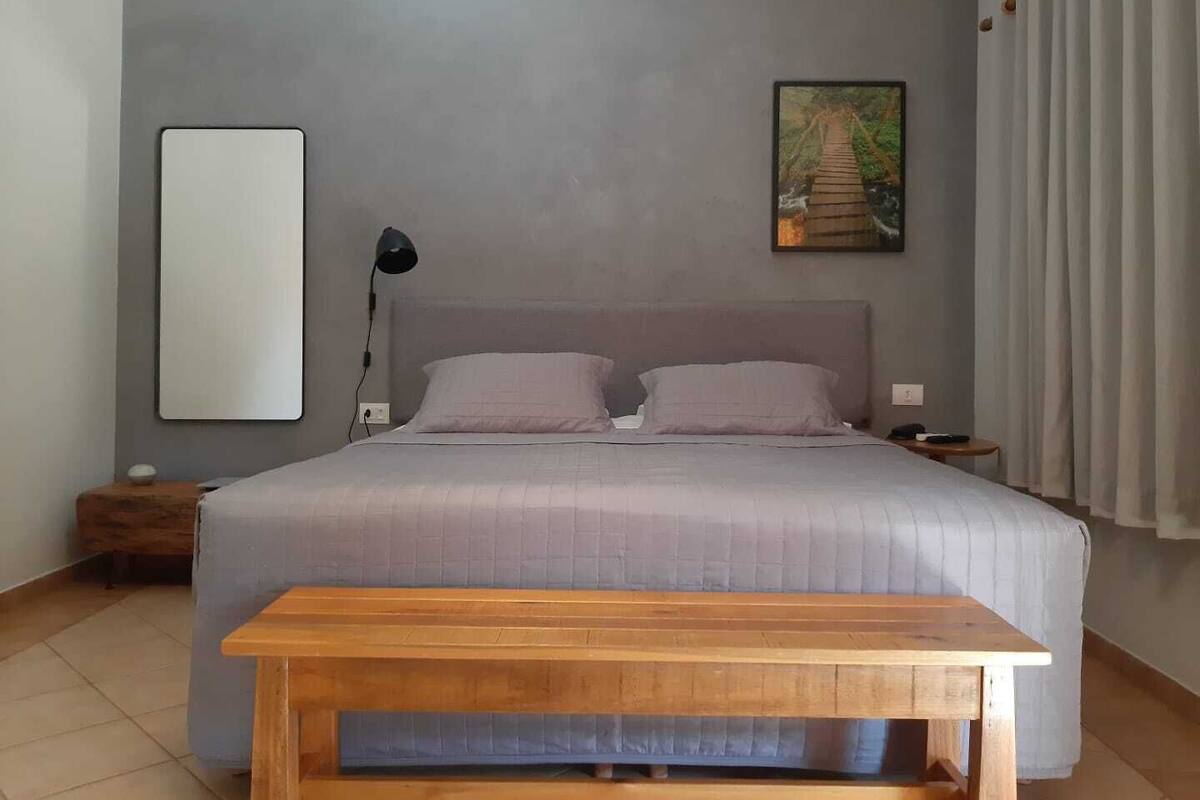 A bedroom with a large bed, a bedside table, a lamp, a bench, and a painting on the wall.