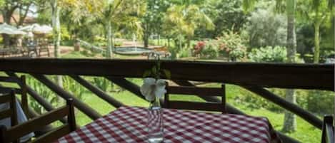 A table with a checkered tablecloth, a vase with flowers, and a view of a lush garden with trees and a fountain.