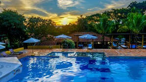 Una piscina con tumbonas y sombrillas, rodeada de palmeras y una estructura de techo con azulejos.