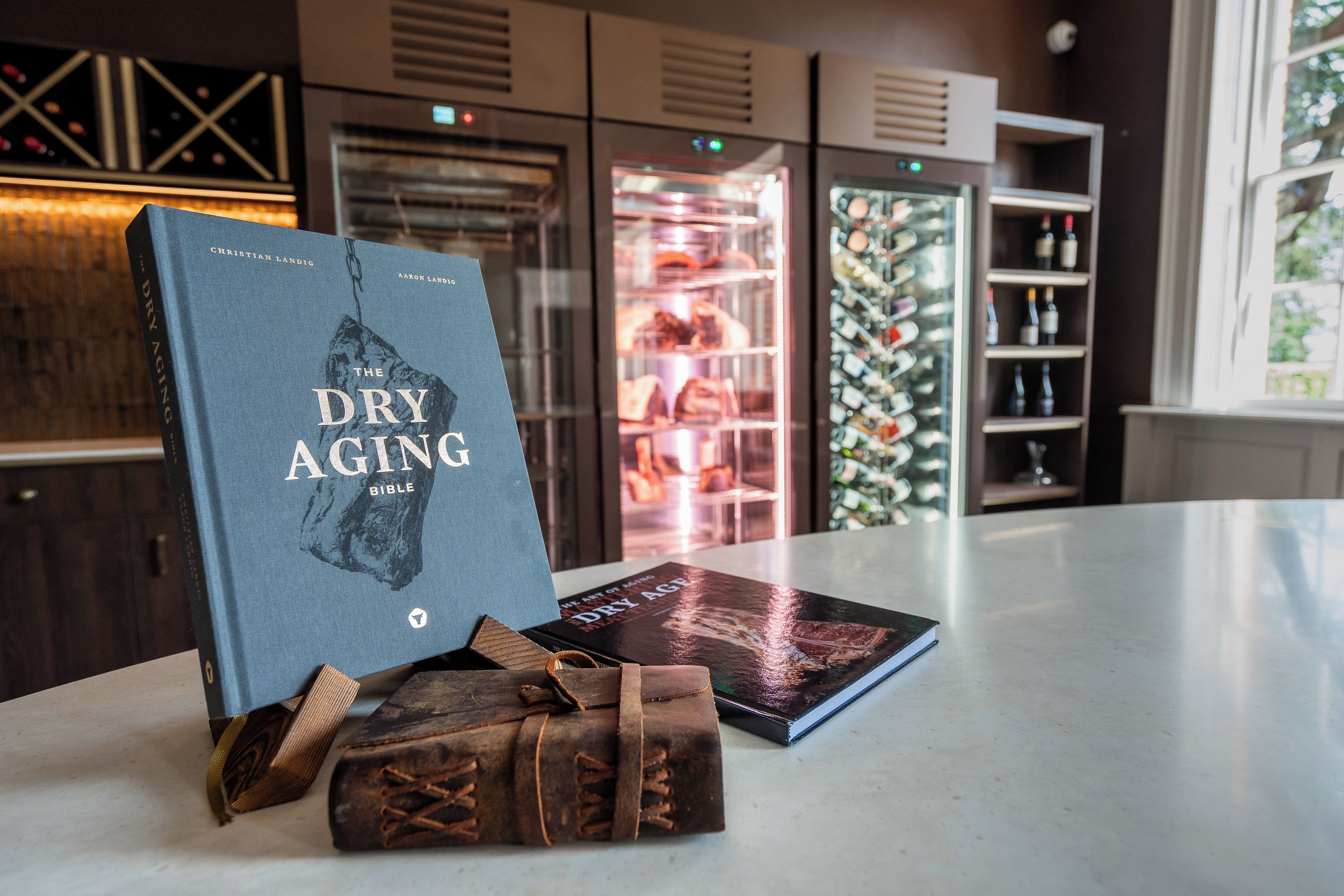 A book titled 'The Dry Aging Bible' on a table with a leather-bound book and a wine bottle.