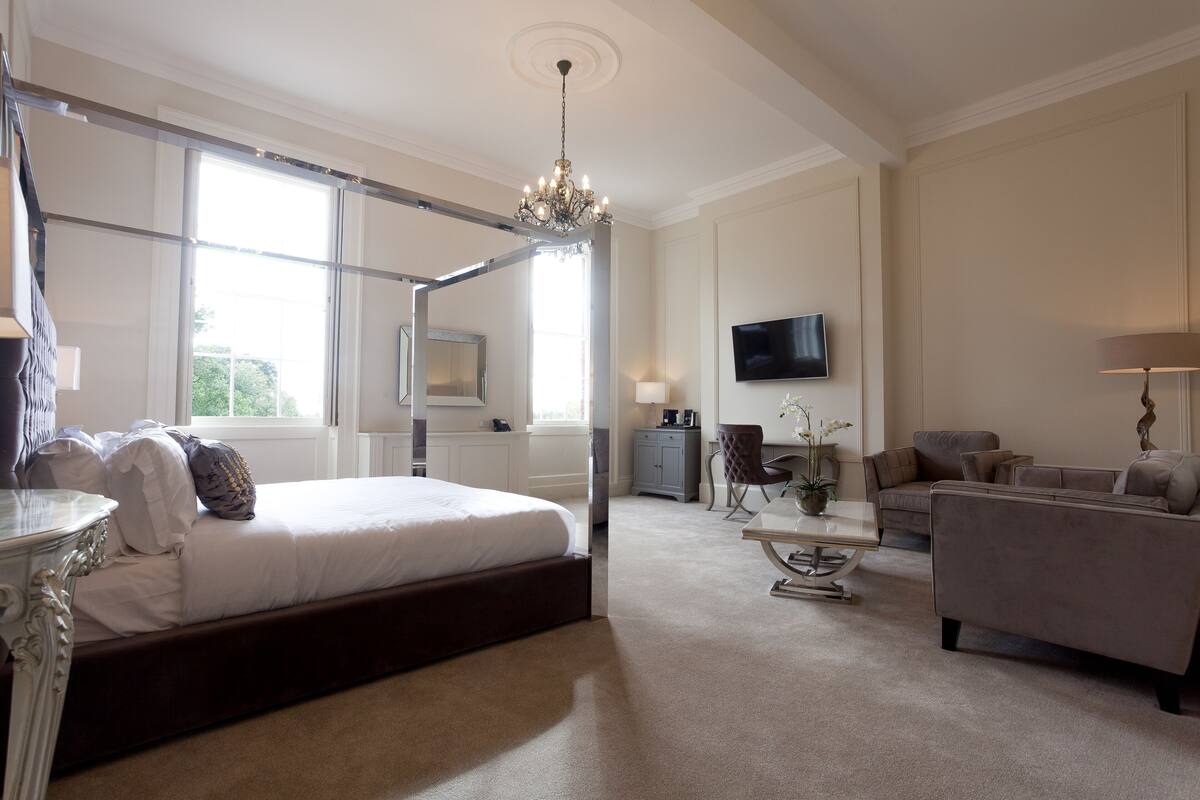 Une chambre spacieuse dotée d’un lit à baldaquin, d’un lustre, d’une télévision et d’un coin salon avec un canapé et un fauteuil.