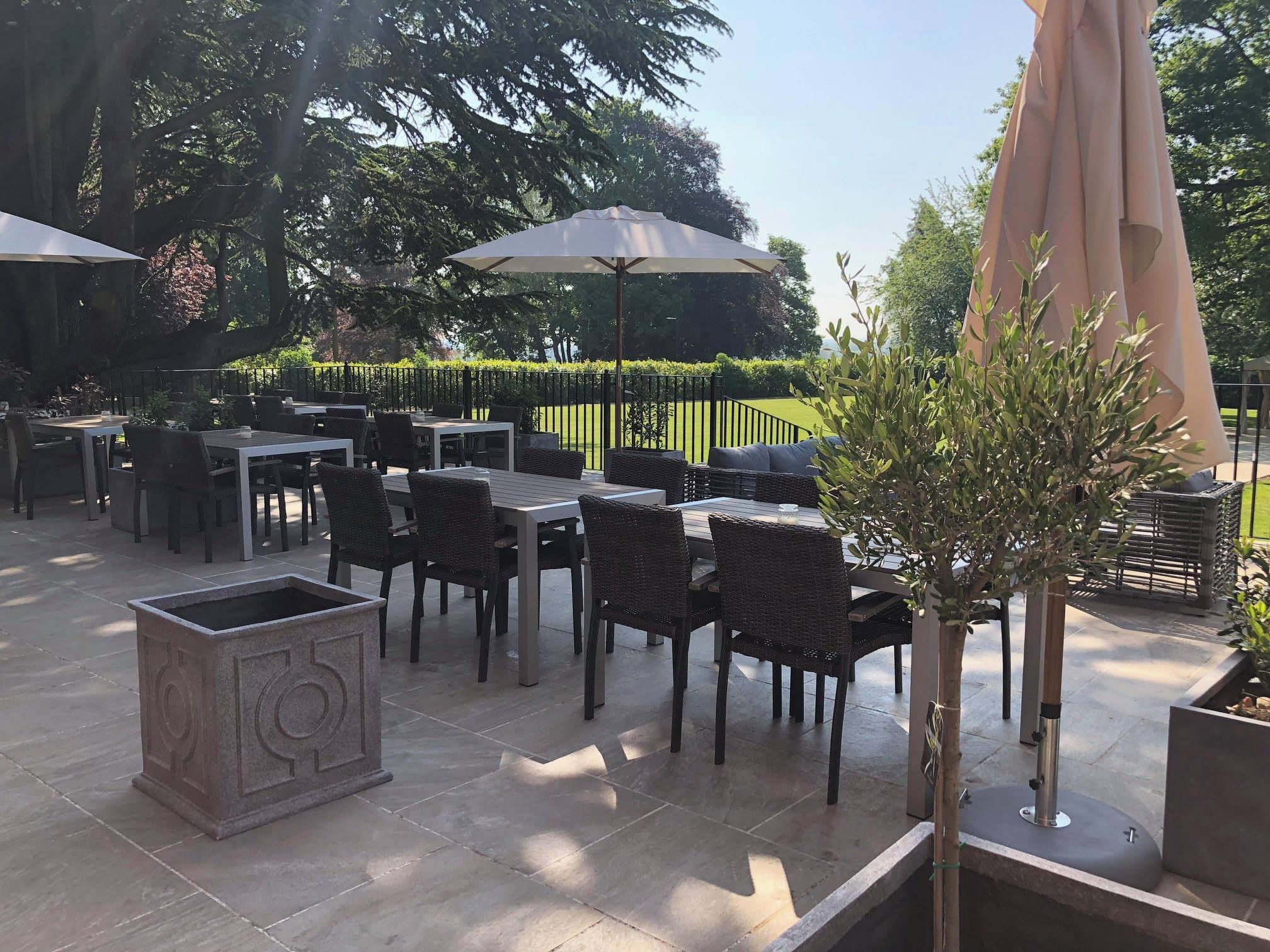 An outdoor patio with tables and chairs, surrounded by greenery and trees.