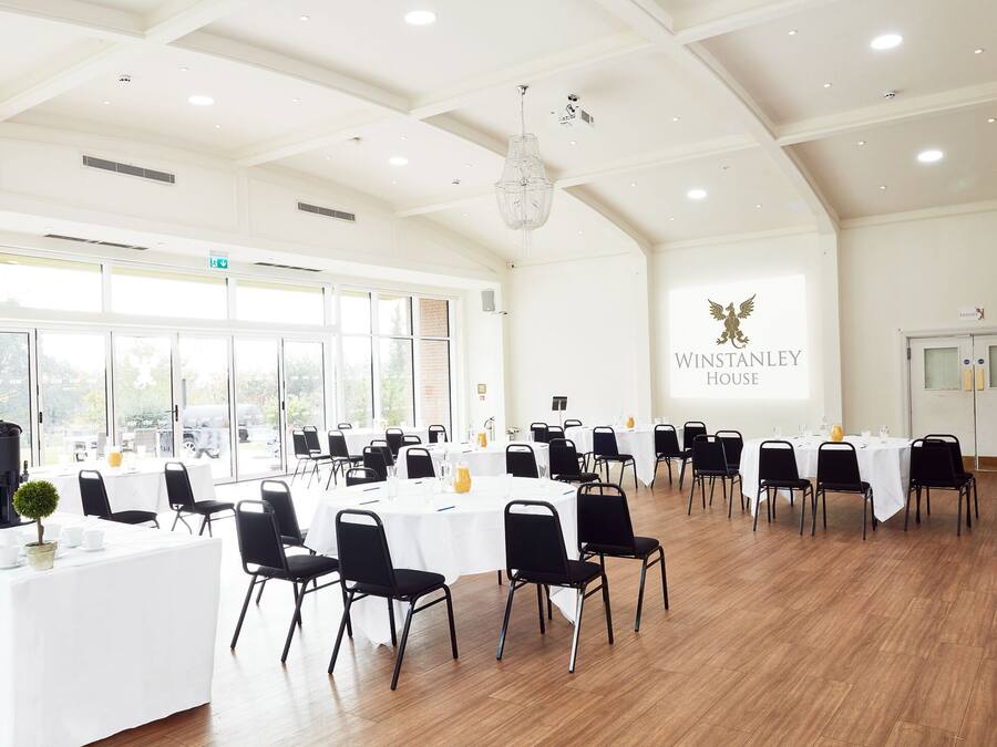A spacious conference room with multiple tables and chairs, large windows, and a chandelier.