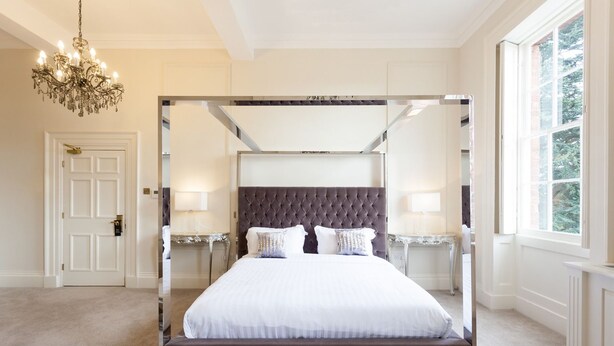 A four-poster canopy bed with a tufted headboard, bedside tables with lamps, and a chandelier. - Winstanley House (Leicester)