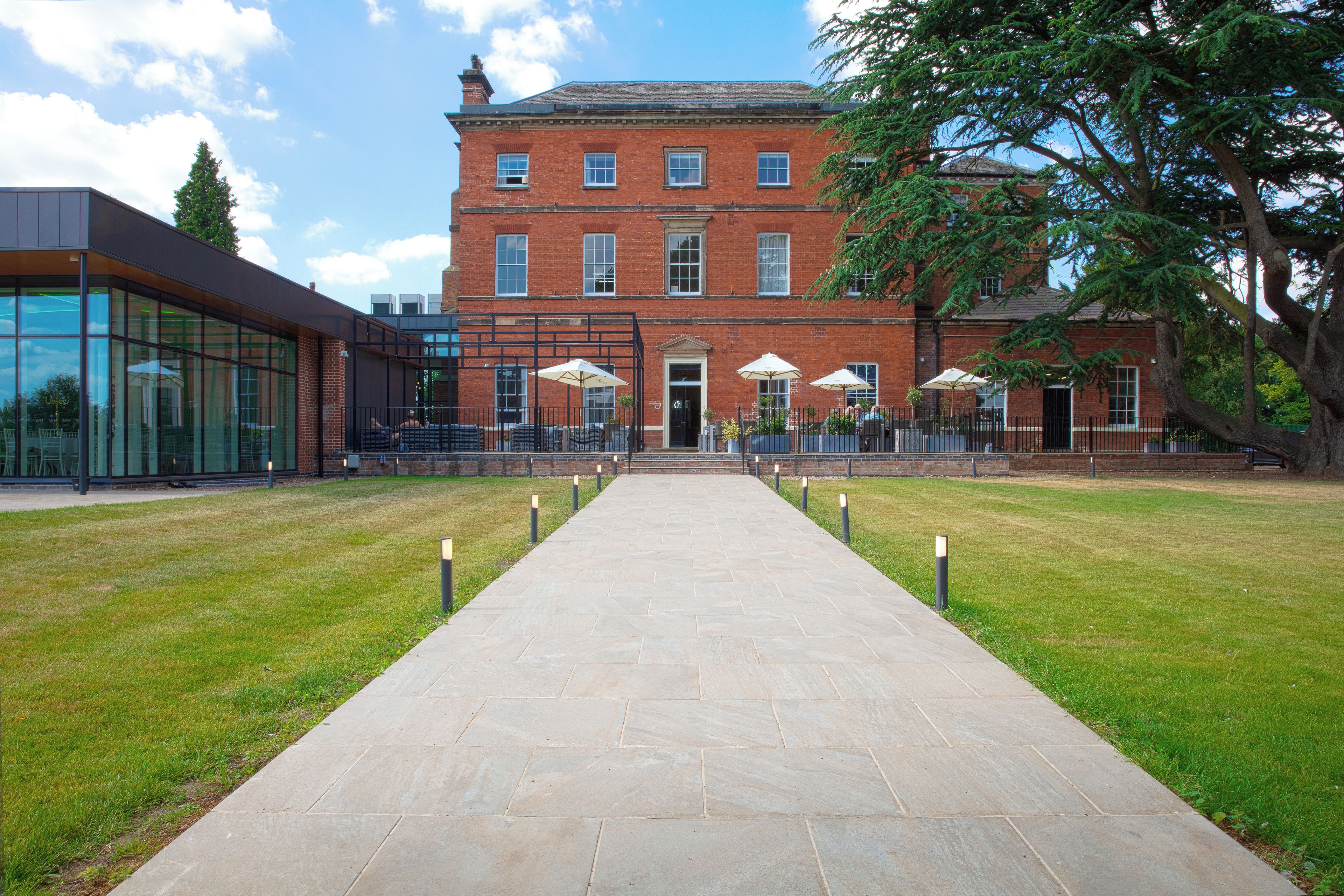 A pathway leading to a building with a brick facade, outdoor seating with umbrellas, and a modern glass structure to the left.