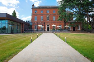 A pathway leading to a building with a brick facade, outdoor seating with umbrellas, and a modern glass structure to the left.