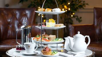 A three-tiered tray of assorted pastries and sandwiches on a table, accompanied by a teapot, cups, and a pitcher, set against a backdrop of a leather couch and decorative lights.