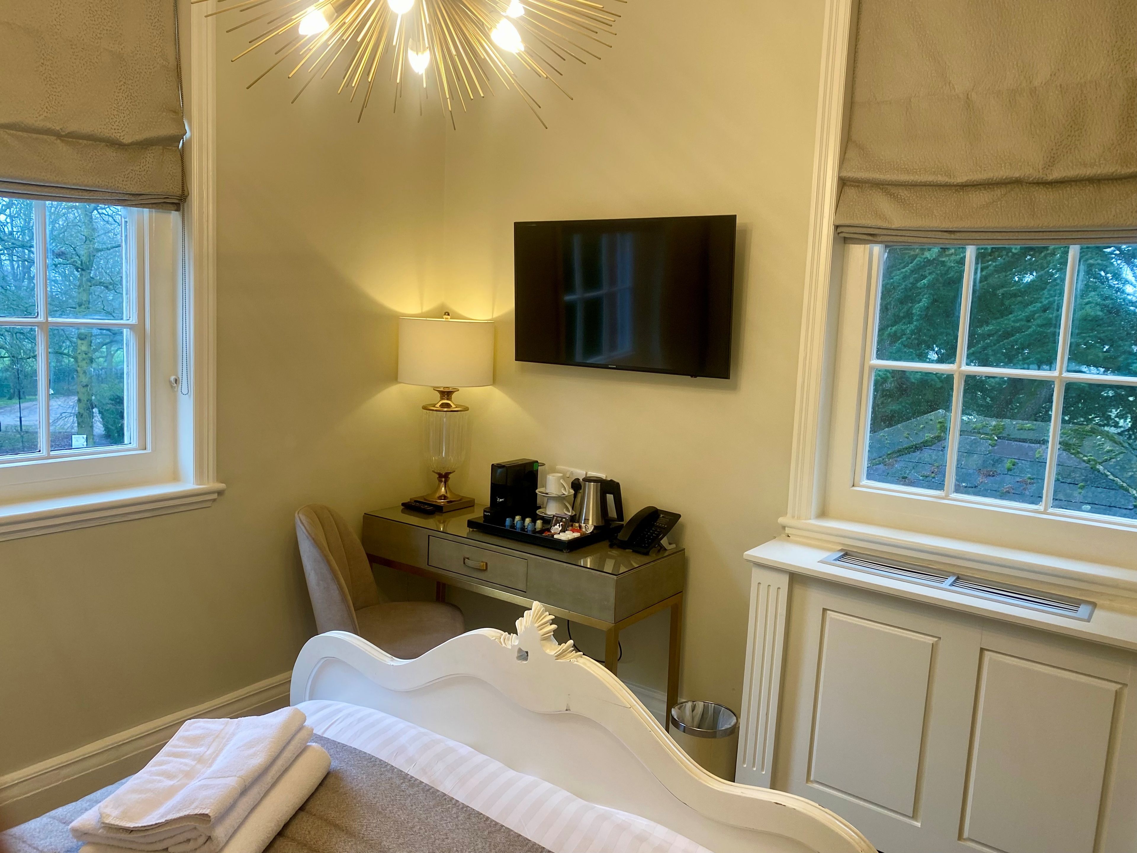 A hotel room with a bed, a desk with a lamp, a television, and a view of trees through the windows.