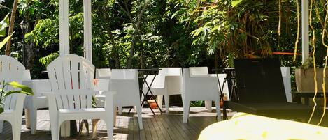 A covered outdoor seating area with white chairs and tables, surrounded by lush greenery.