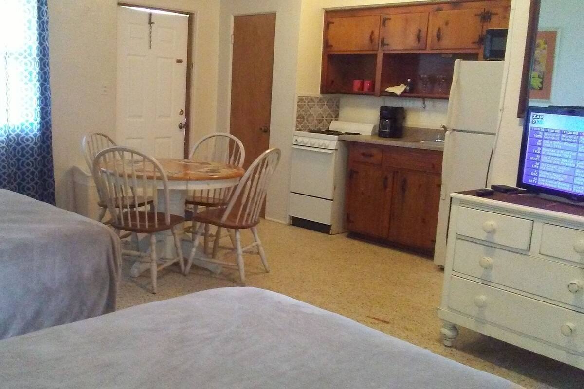 A small, compact room with two single beds, a dining table with four chairs, a kitchenette with a stove and refrigerator, and a TV on a dresser.