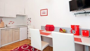 A modern kitchen with white cabinets, a wooden ceiling, a red pendant light, a dining area with white chairs, a red coffee maker, and a flat-screen TV mounted on the wall. - Apartments Neva (Pula)