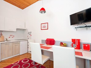 A modern kitchen with white cabinets, a wooden ceiling, a red pendant light, a dining area with white chairs, a red coffee maker, and a flat-screen TV mounted on the wall. - Apartments Neva (Pula)