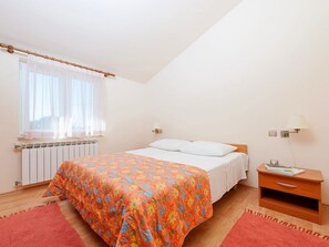 A bedroom with a bed, a nightstand, a lamp, a window with sheer curtains, and a radiator. - Apartments Neva (Pula)