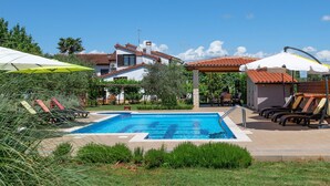 A swimming pool with lounge chairs and umbrellas, surrounded by greenery and a building with a tiled roof. - Apartments Neva (Pula)