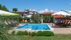 A swimming pool with lounge chairs and umbrellas, surrounded by greenery and a building with a tiled roof. - Apartments Neva (Pula)