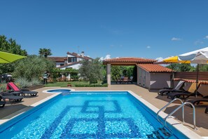 Una piscina con sillas de descanso y sombrillas, rodeada de edificios y follaje.