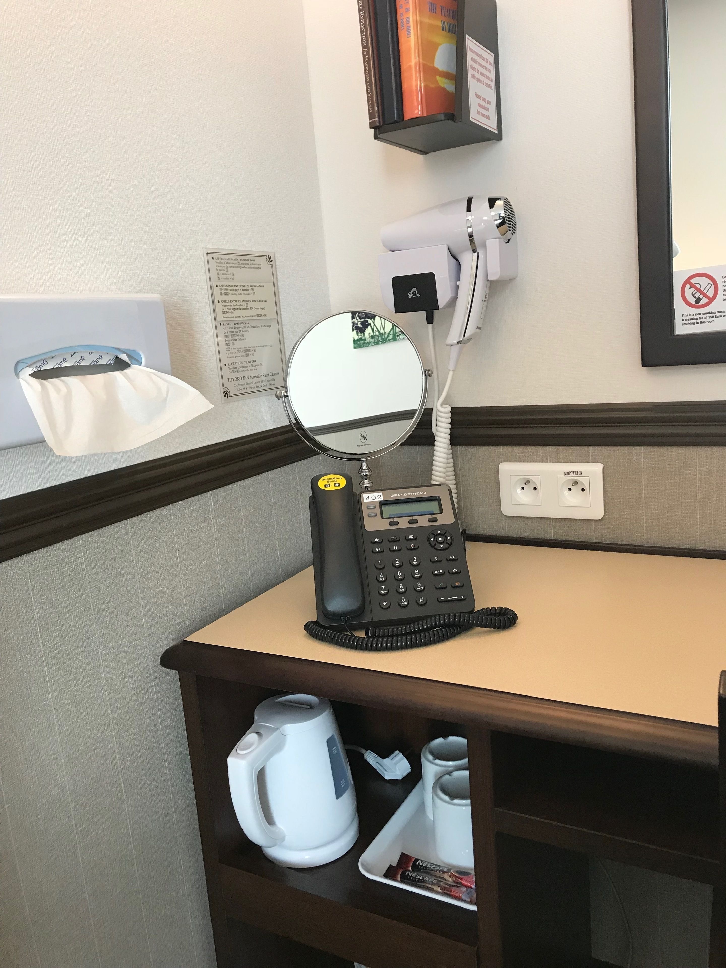 Mesa de quarto de hotel com telefone, espelho, secador de cabelo, caixa de lenços e máquina de café.