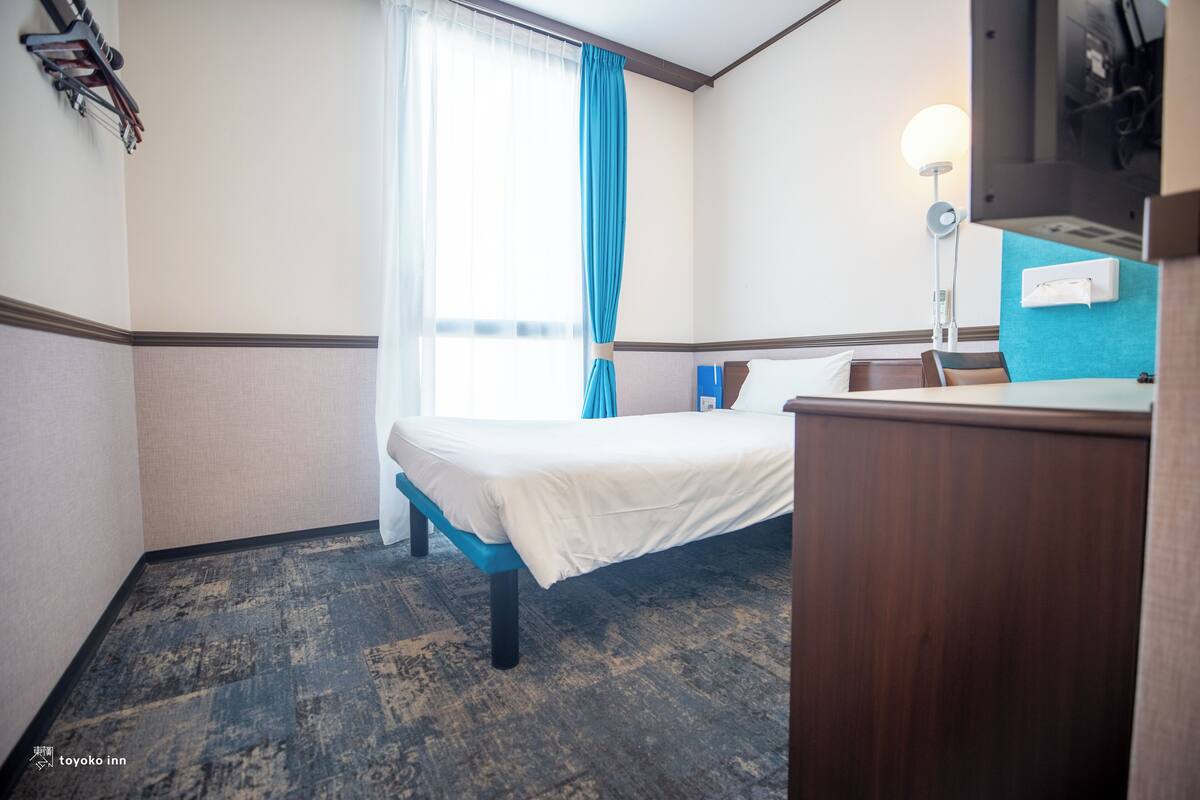 Camera d'albergo con un letto, una scrivania, una sedia, uno specchio, una lampada e una finestra con tende.