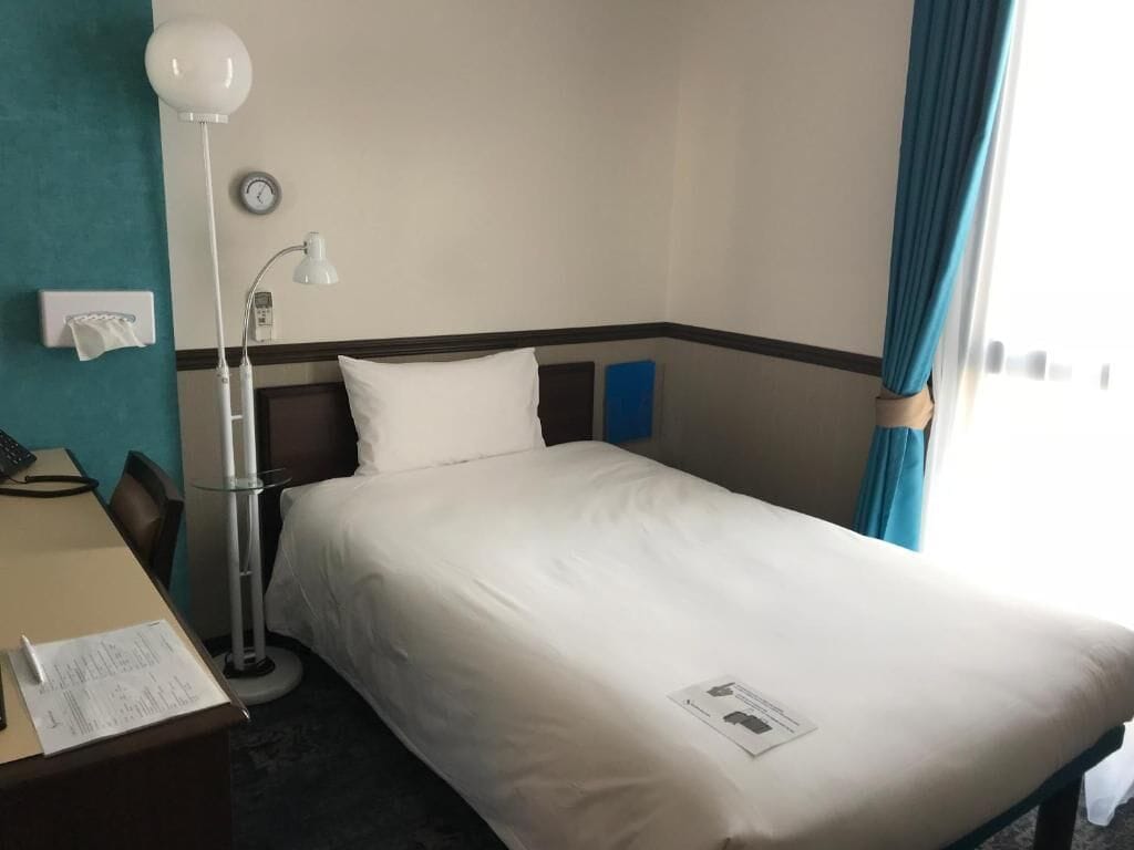 Una camera d'albergo con un letto, una scrivania con lampada, una sveglia al muro e una finestra con tende.