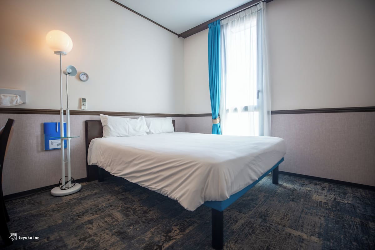 Una camera d'hotel con un letto, una lampada da terra, una sveglia a parete e una finestra con tende.