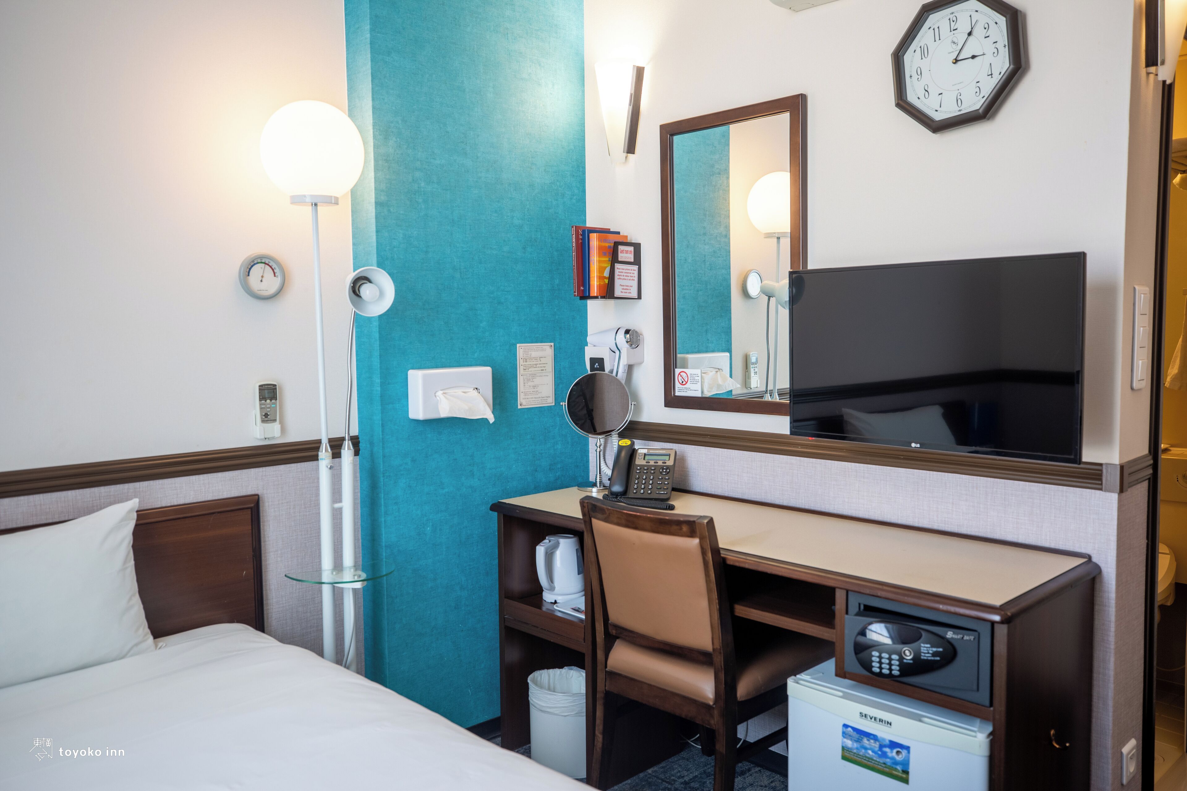 Une chambre d’hôtel comprenant un lit, un bureau avec une chaise, une lampe de bureau, une horloge, un miroir et un mini-bar.