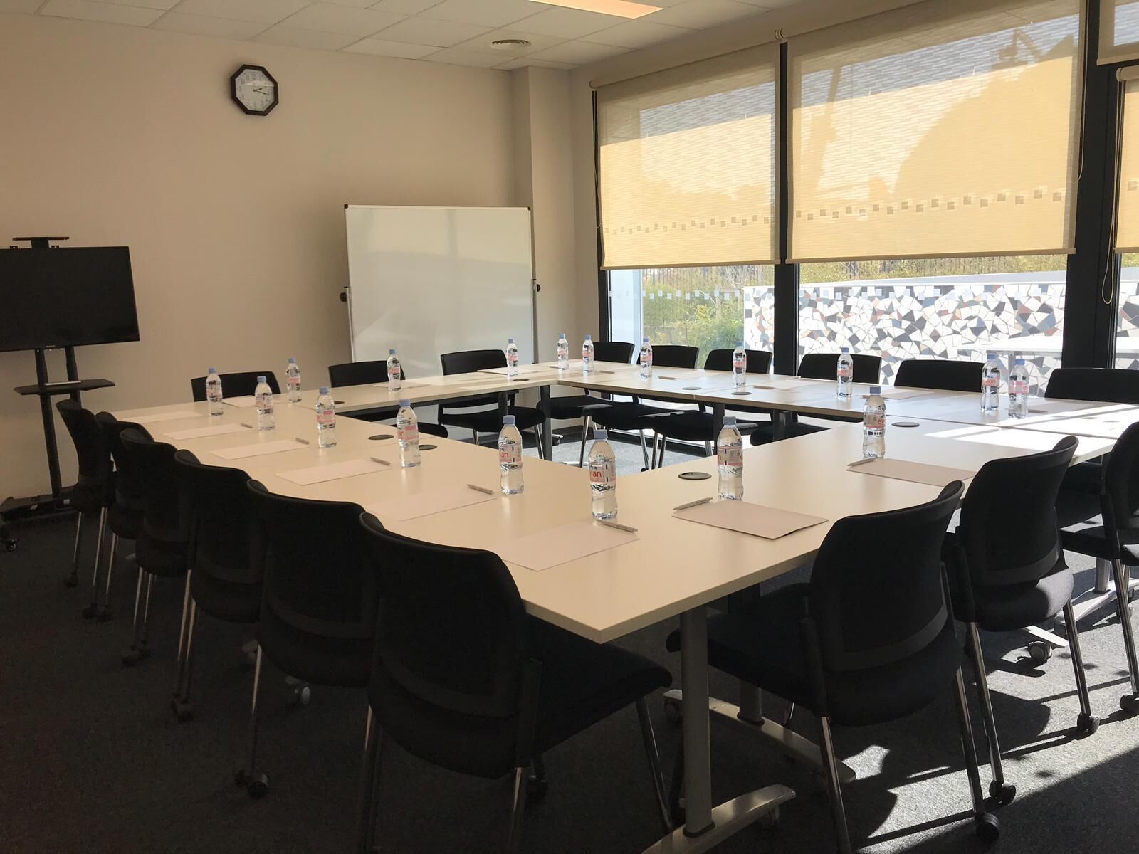 A conference room with a long table, black chairs, water bottles, a whiteboard, and a clock on the wall.
