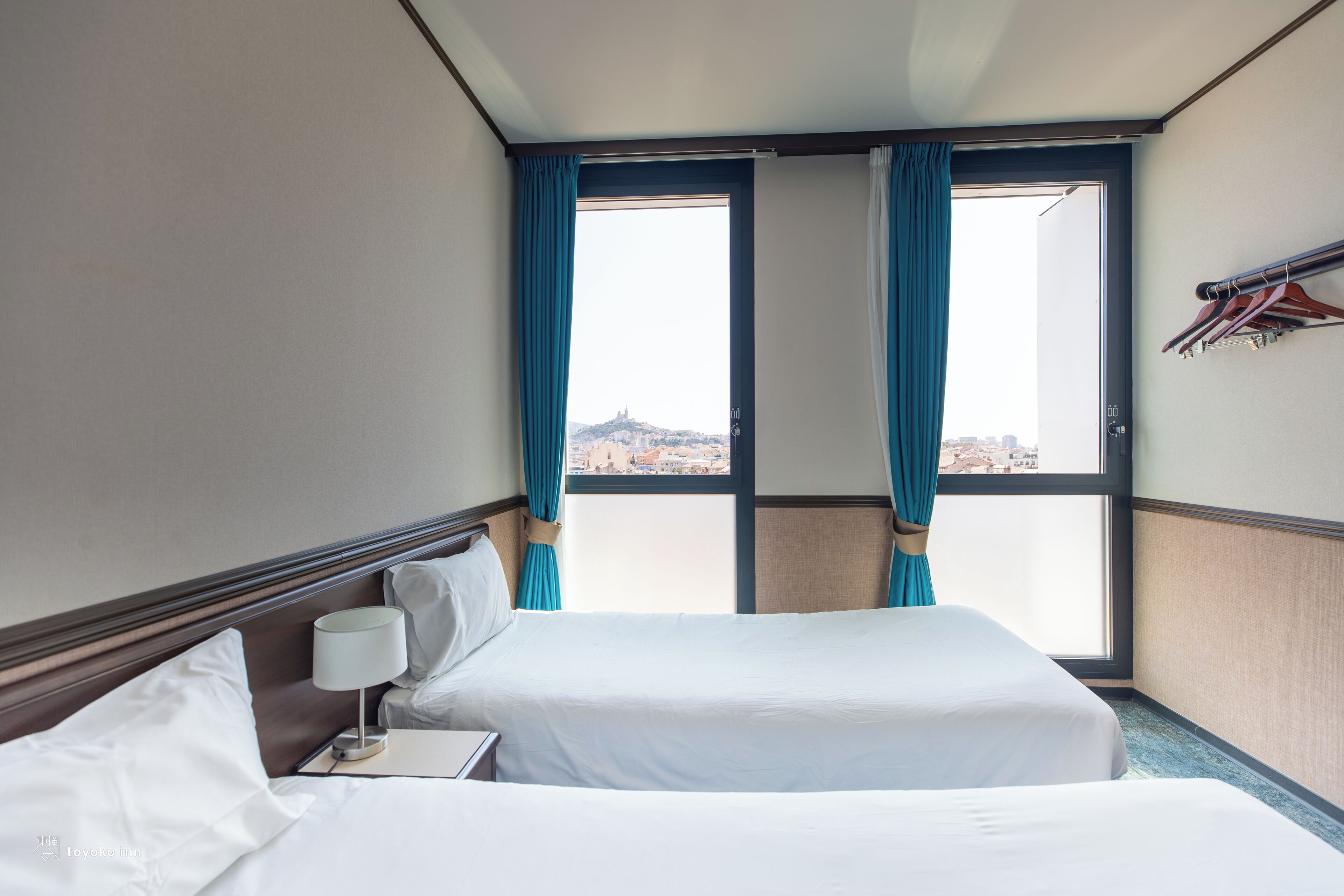 Ein Hotelzimmer mit zwei Betten, einem Nachttisch, einer Lampe und Blick auf die Stadt durch große Fenster mit blauen Vorhängen.