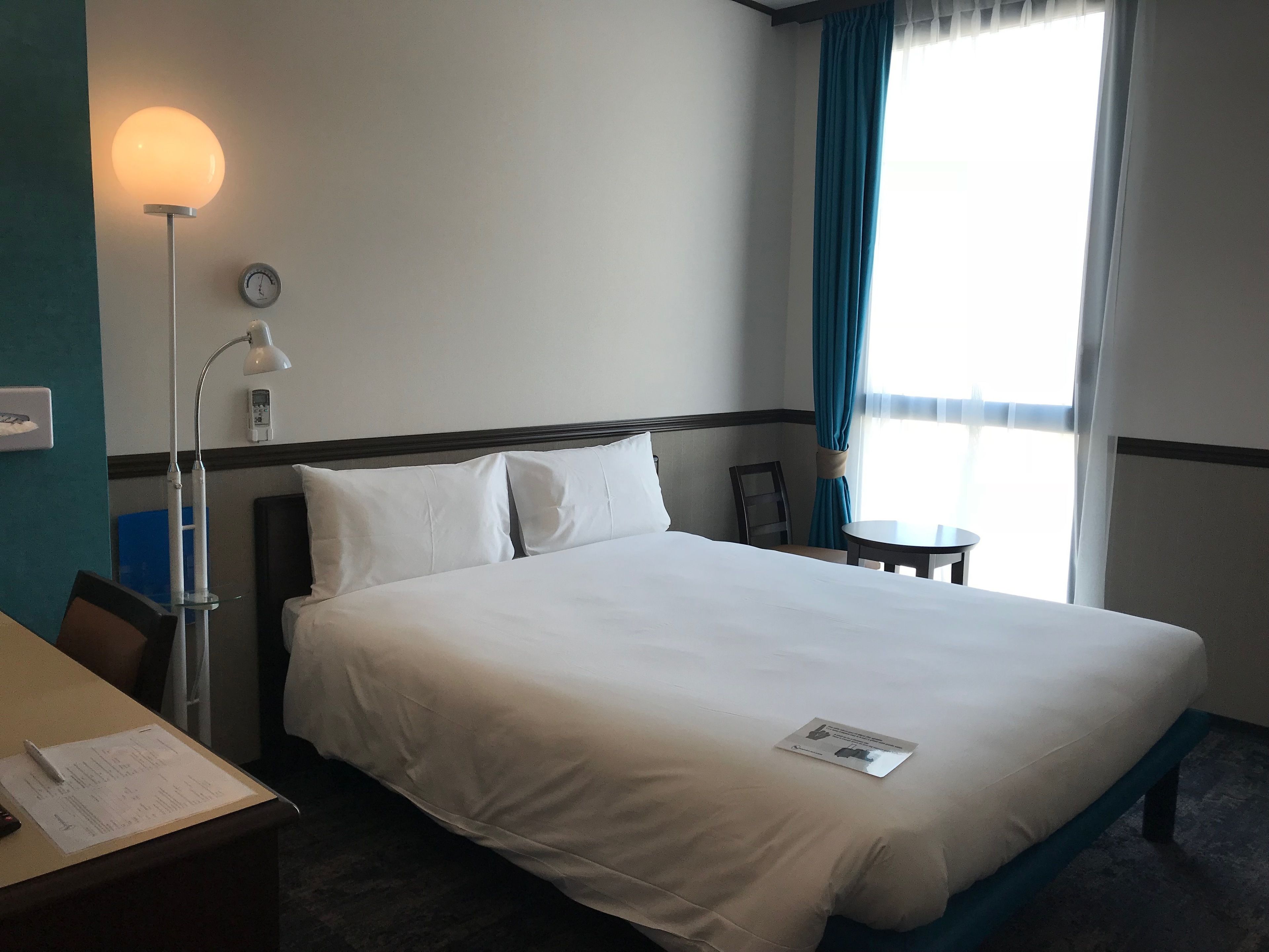 Habitación de hotel con cama, escritorio, lámpara, reloj y ventana con cortinas.