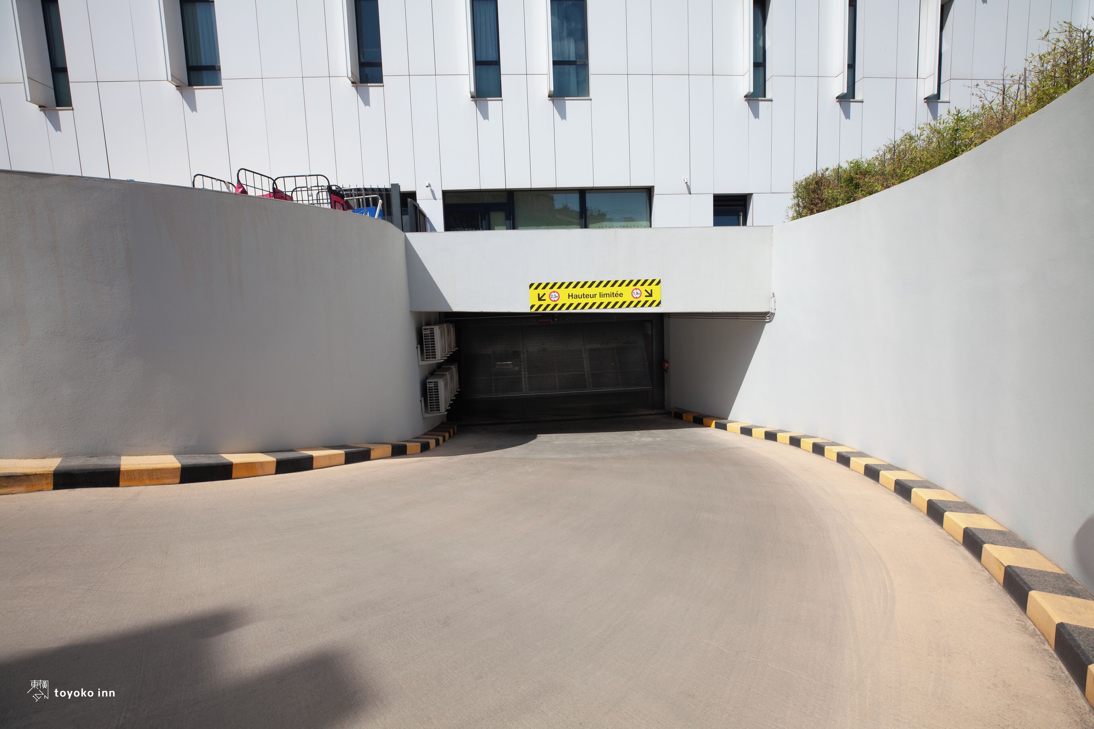 Un área de estacionamiento con un letrero de precaución amarillo, un edificio con varias ventanas y una borda con marcas negras y amarillas.