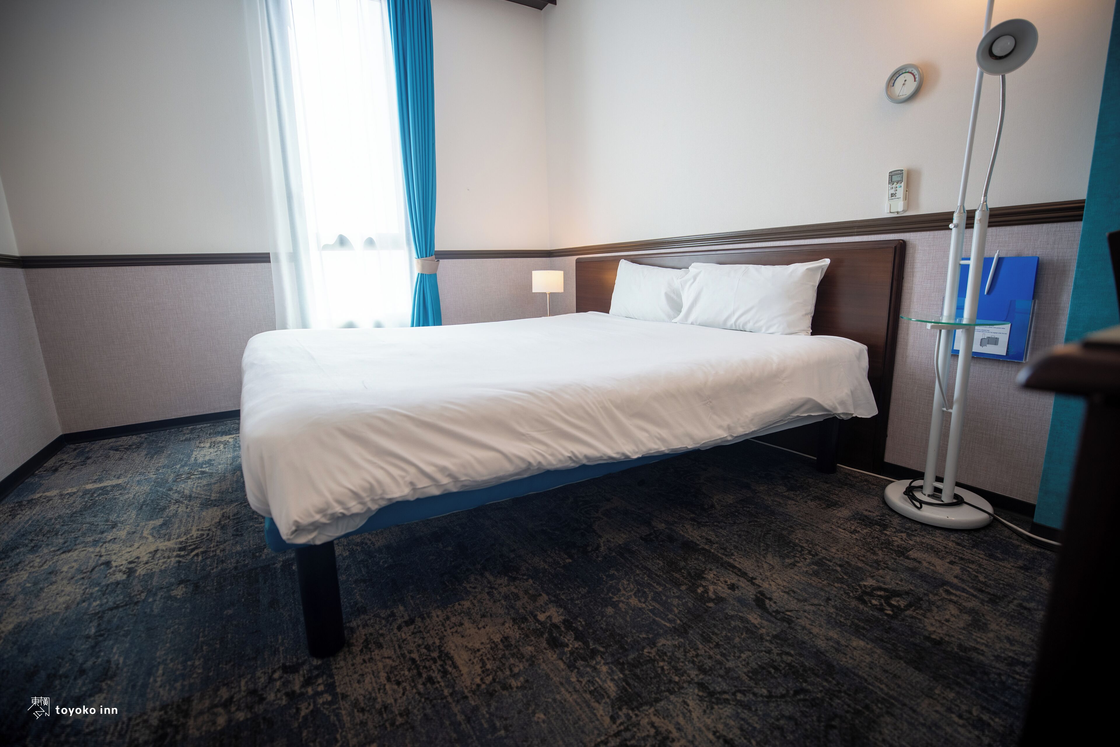 A hotel room with a bed, a bedside table, a lamp, a clock, and a blue curtain.