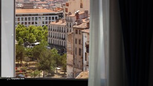 Vista desde una ventana con cortinas, que da a un paisaje urbano con edificios y una colina con una estructura destacada.