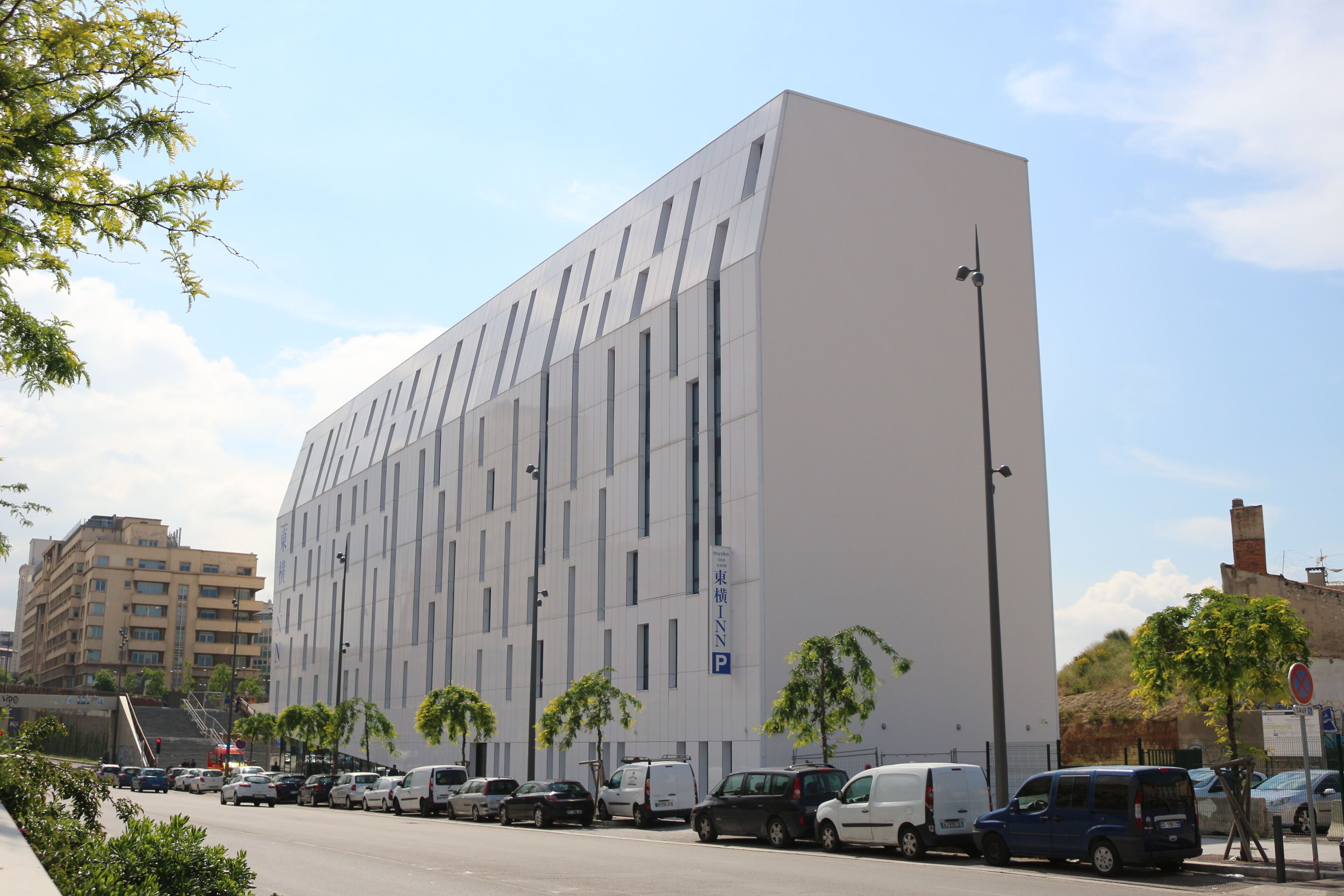 A modern building with a flat facade, multiple windows, and a parking area with various vehicles.