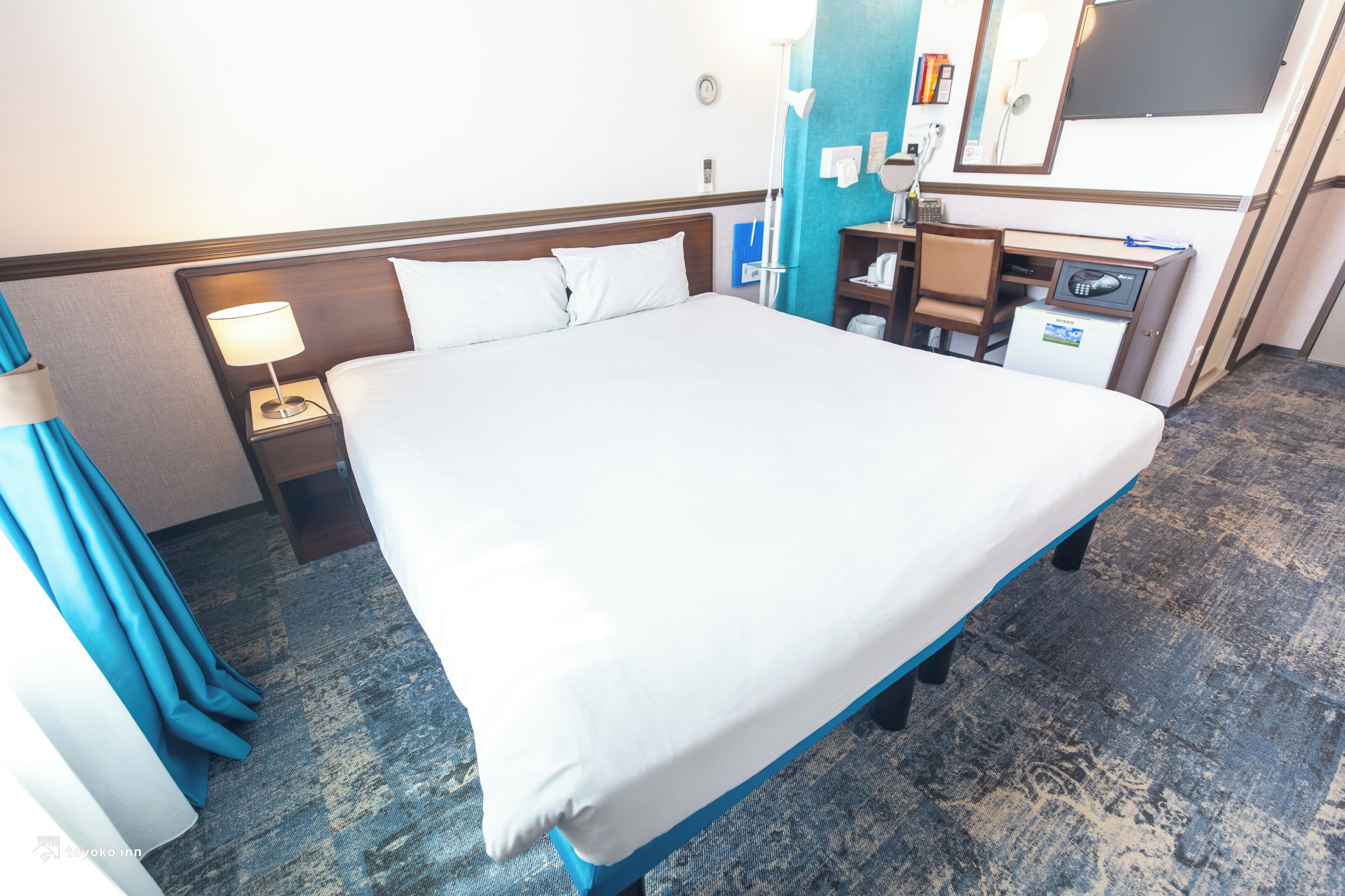Quarto de hotel com uma cama grande, uma escrivaninha, uma cadeira, um pequeno frigorífico e uma mesa de cabeceira com uma luminária.
