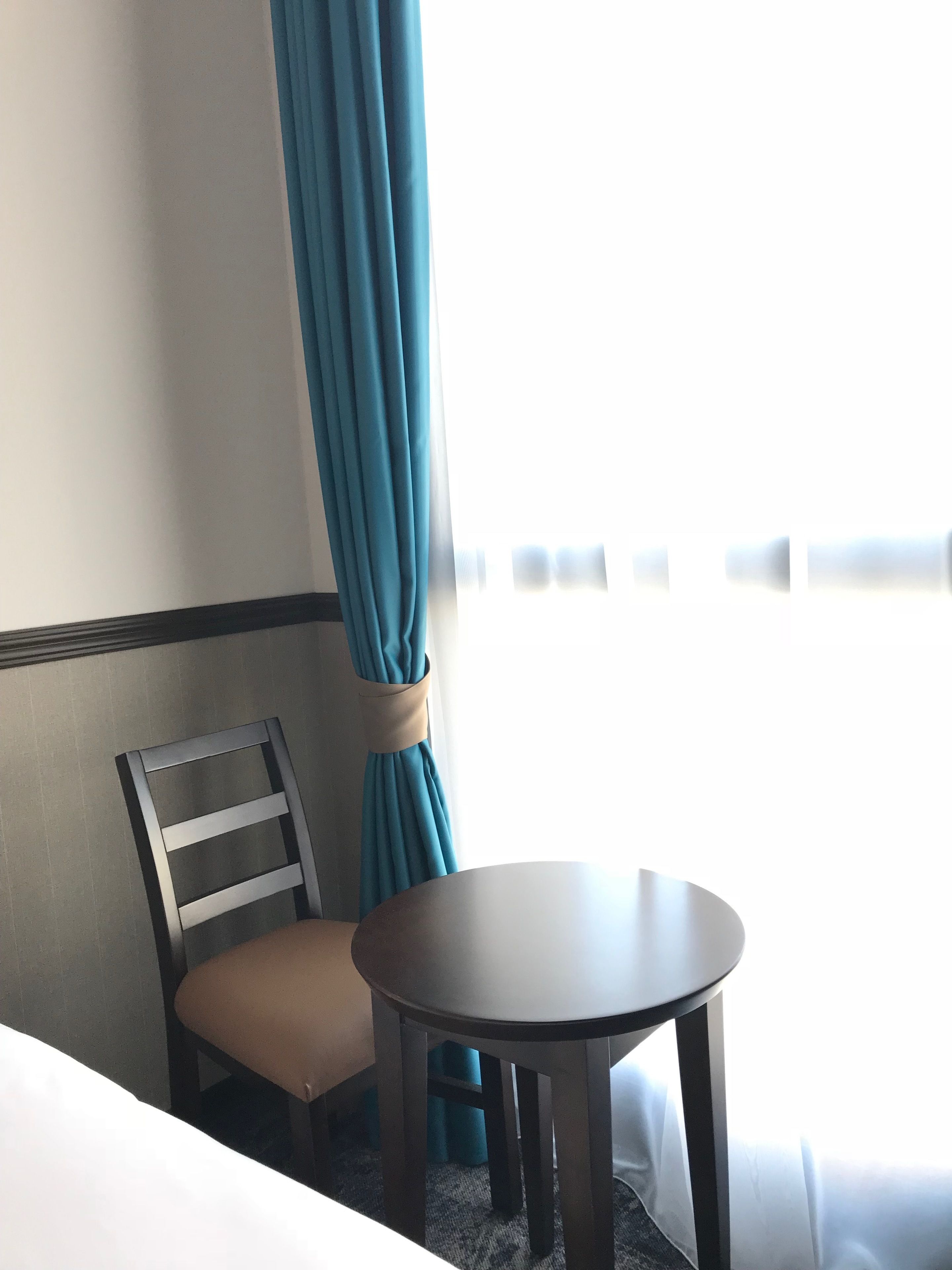 Una habitación de hotel con una cama individual, una mesita redonda pequeña, una silla y un ventanal con cortinas.