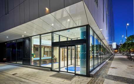 A modern hotel entrance with glass doors, a reception desk, and a blue carpet inside.