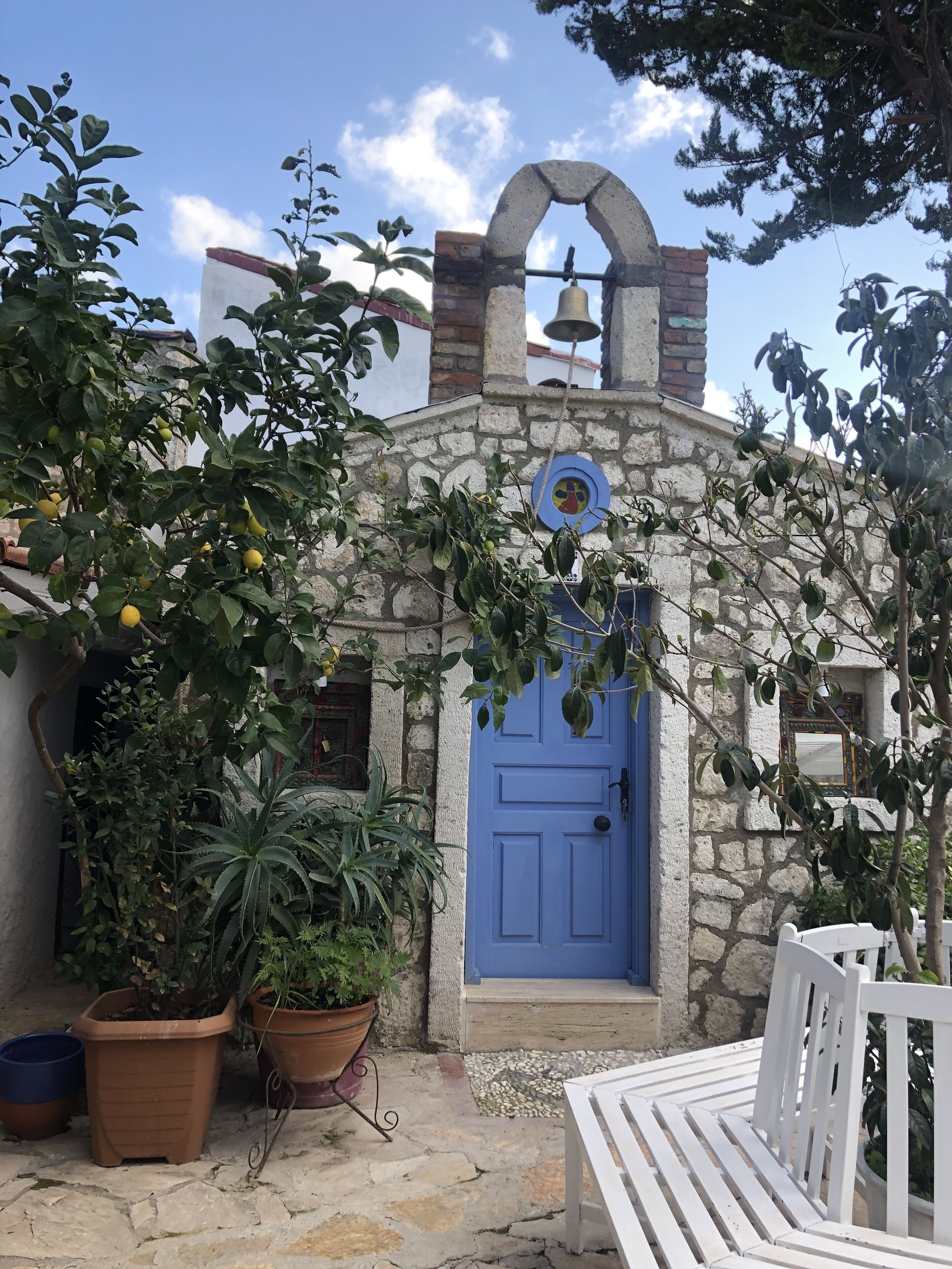 Un bâtiment en pierre avec une porte bleue, des plantes en pot et une cloche au-dessus de l’entrée.