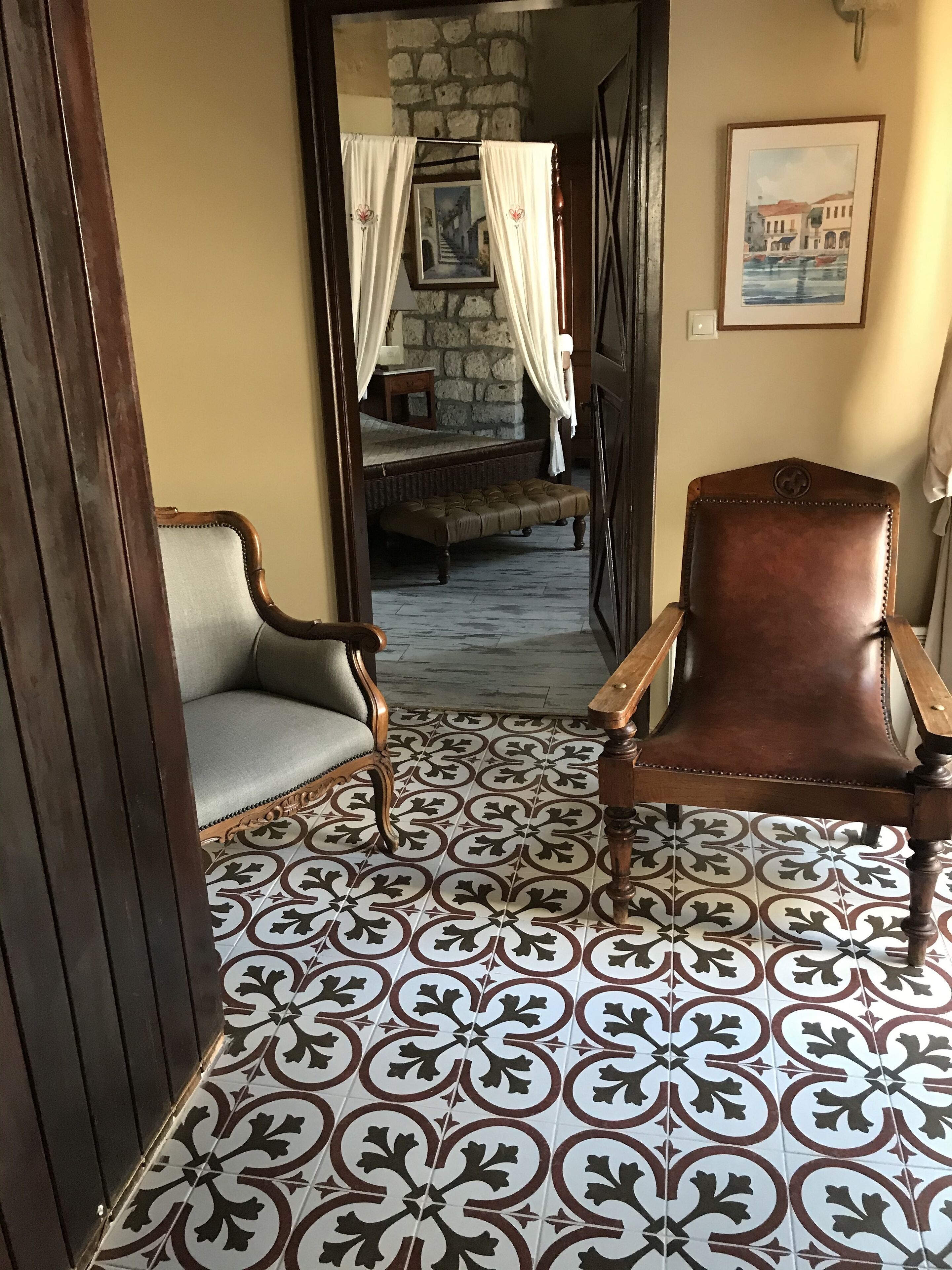A room with a patterned floor, a wooden chair, a cushioned bench, and a bed with a stone wall backdrop.
