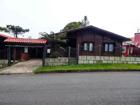 Uma casa de madeira de dois andares com telhado de telhas vermelhas, um caminho de pedra que leva à entrada e um portão de metal cercando a propriedade.
